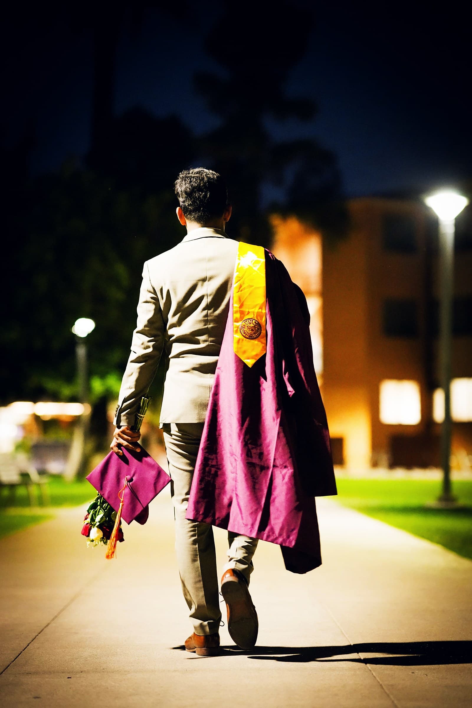 Uday Kumar M walking along a lit campus path at night, carrying his graduation gown, cap, and roses
